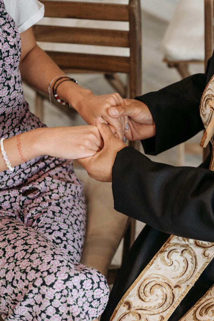 A priest and a woman hold hands in a gesture of support and connection indoors.